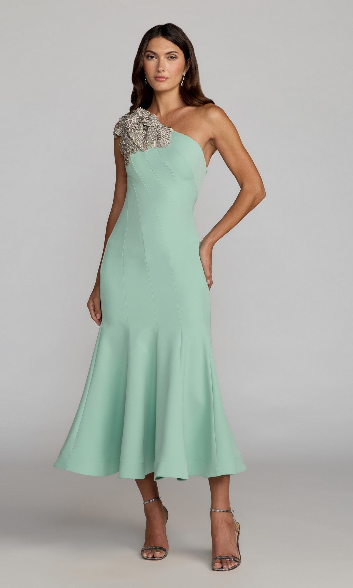 A woman wearing the Mac Duggal 13221 tea-length party dress in sage, a crepe one-shoulder design featuring a large embellished floral applique at the shoulder, stands against a plain gray background.